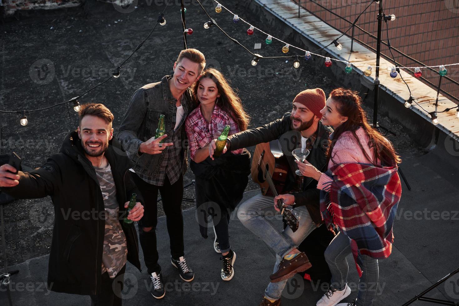 Captures the moment. Top view of a young friends that take selfie while have fun at the roof with decorative colorful light bulbs photo