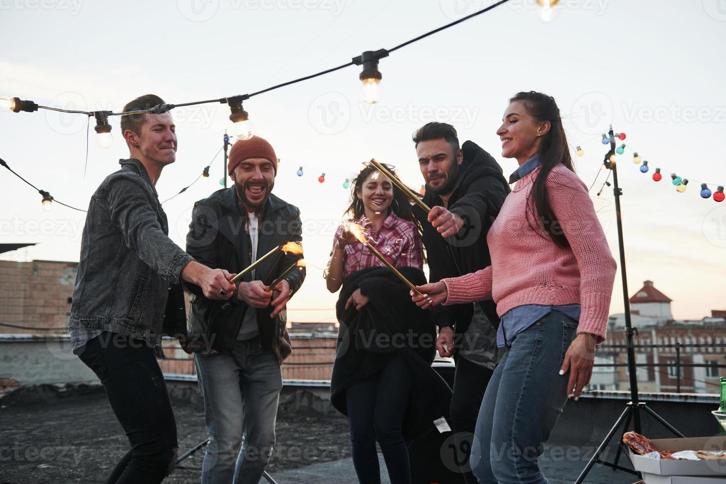 These people are happy to celebrate together. Playing with sparklers on the rooftop. Group of young beautiful friends photo