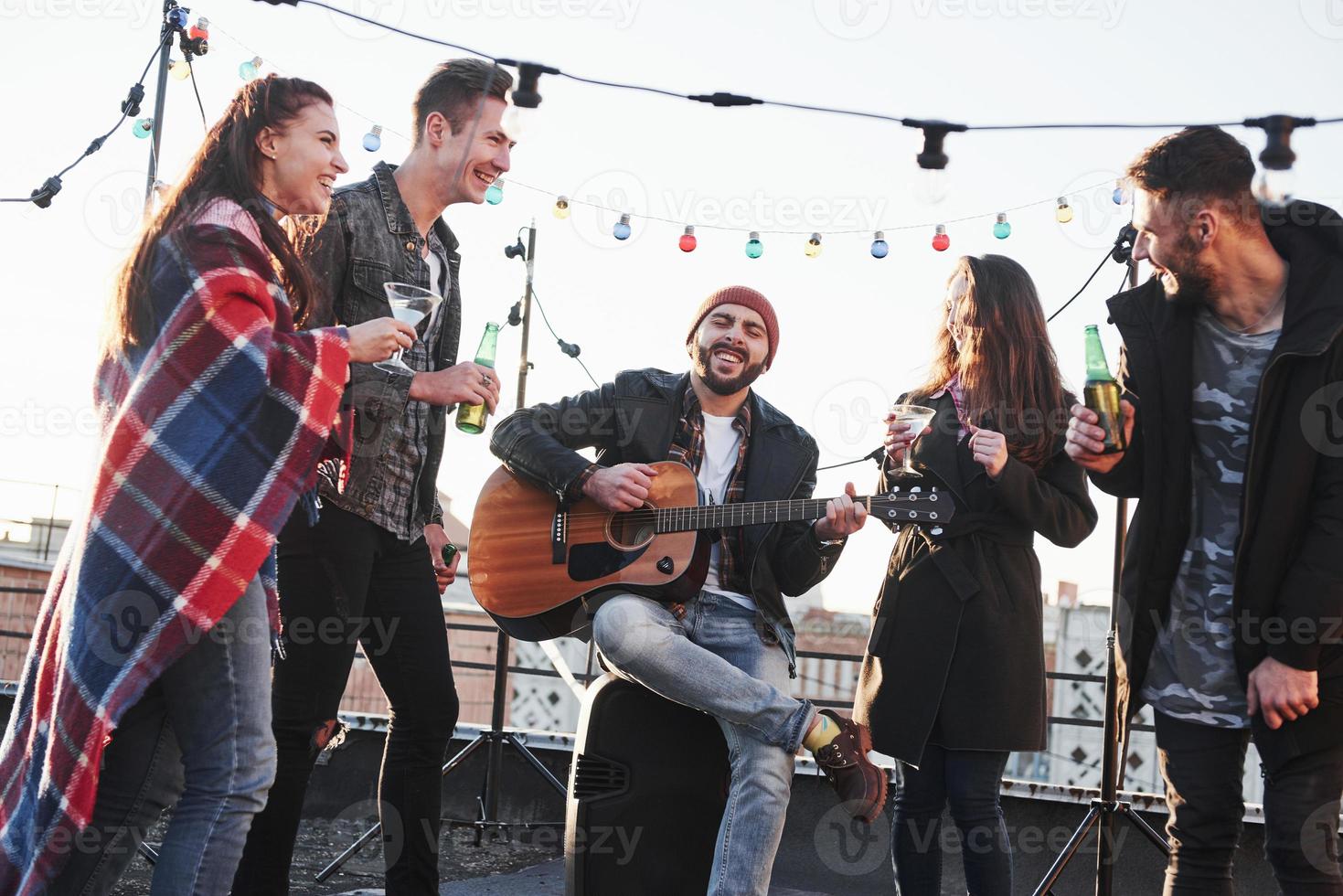el hombre barbudo hace sonreír a todos con su canto. Cinco jóvenes amigos tienen una fiesta con cerveza y guitarra en la azotea. foto