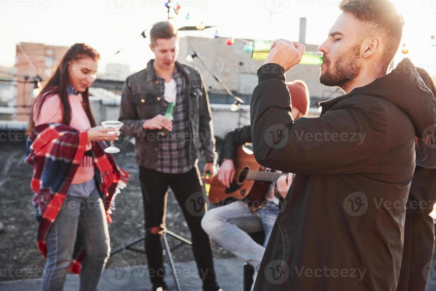 Stylish guy drinks beer. Rooftop party. Friends with alcohol having good time. Guy playing guitar photo