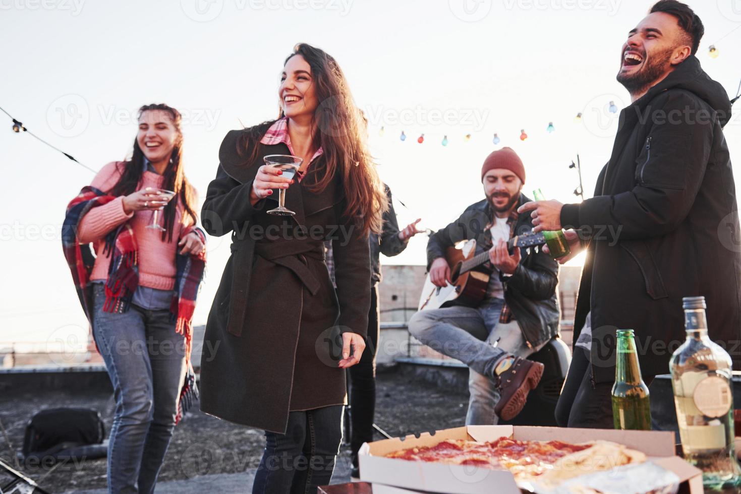 Joking around. Cheerful young people smiling and drinking at the rooftop. Pizza and alcohol on the table. Guitar player photo