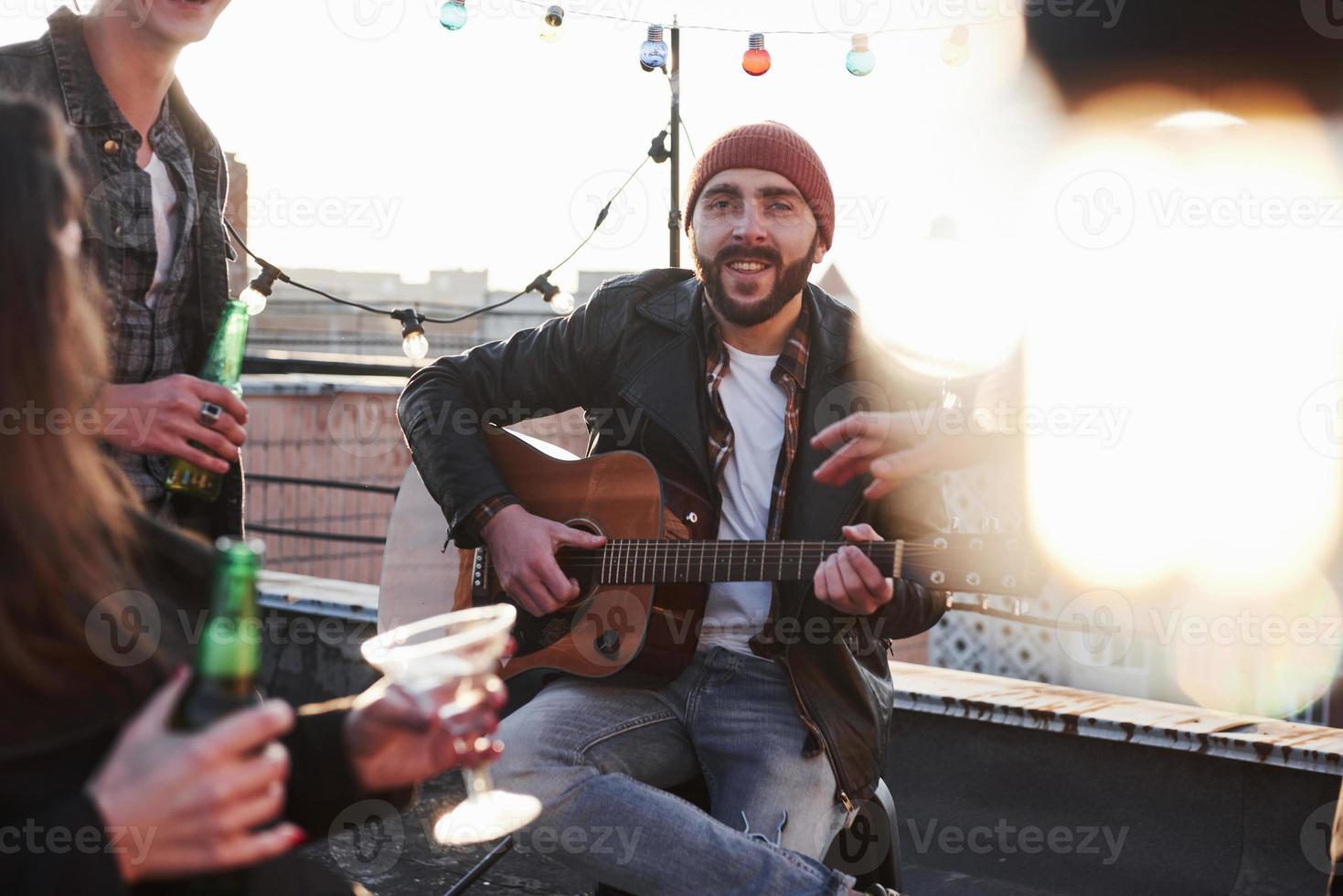 Bearded guitarist smiles into the camera. Three friends with alcohol and acoustic guitar. Blurred bulb in right side of photo