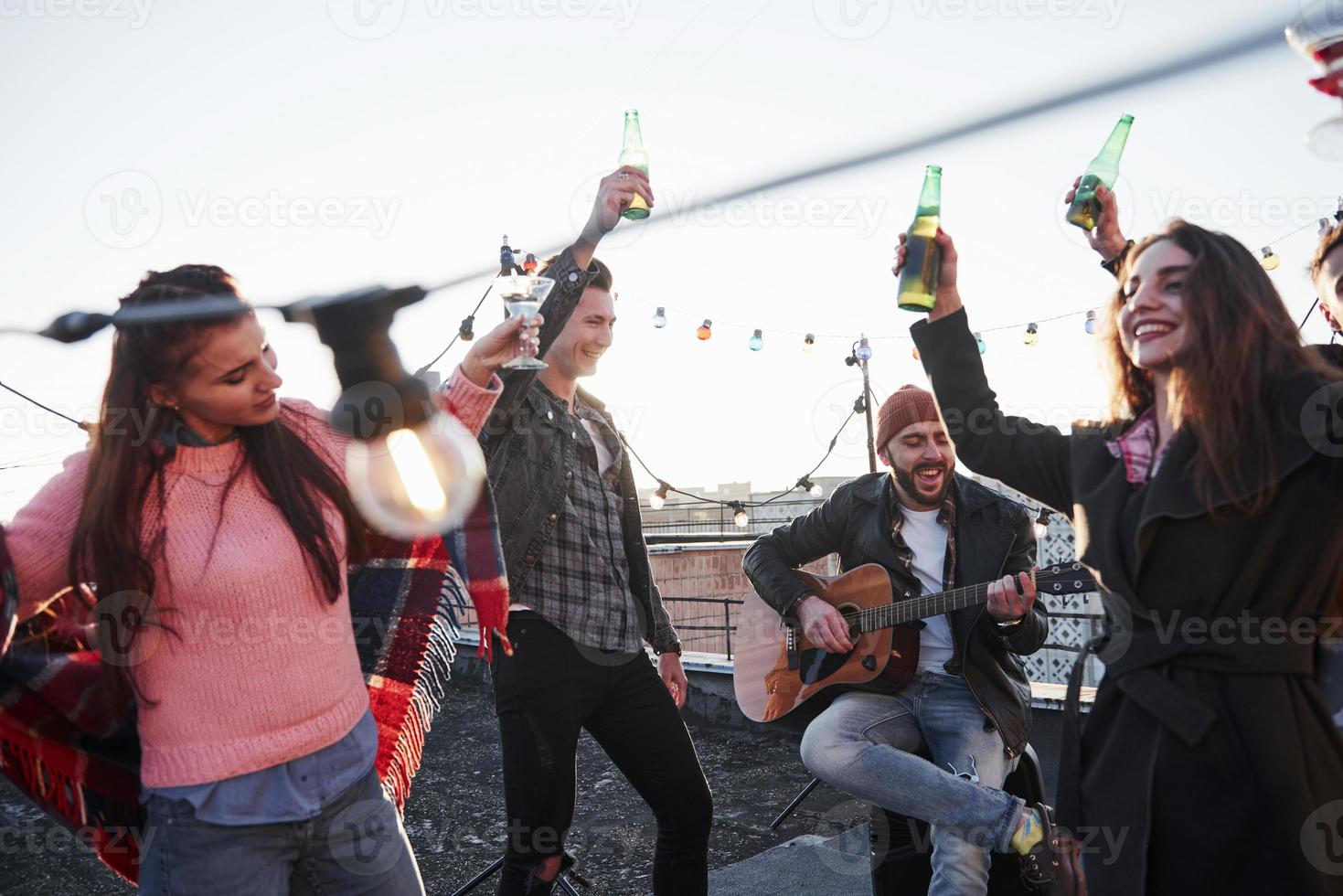 Bottles up and sing. Group of young people having celebration at a rooftop with some alcohol and guitar playing photo