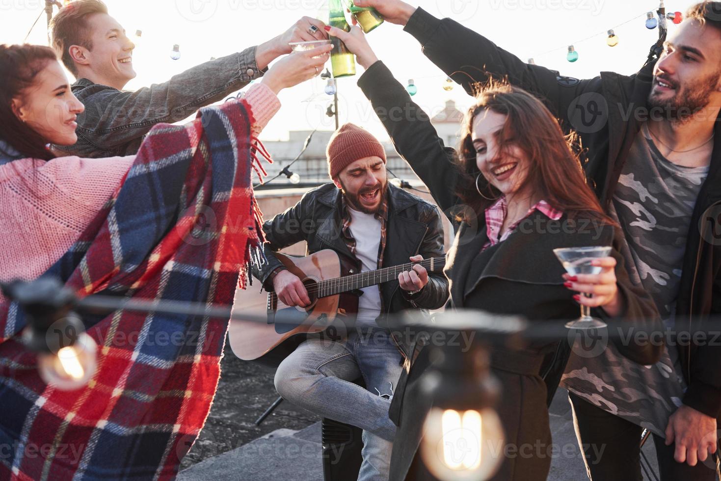 hermoso chico canta mientras amigos toman una copa. Grupo de jóvenes celebrando en una azotea con algo de alcohol y tocar la guitarra foto