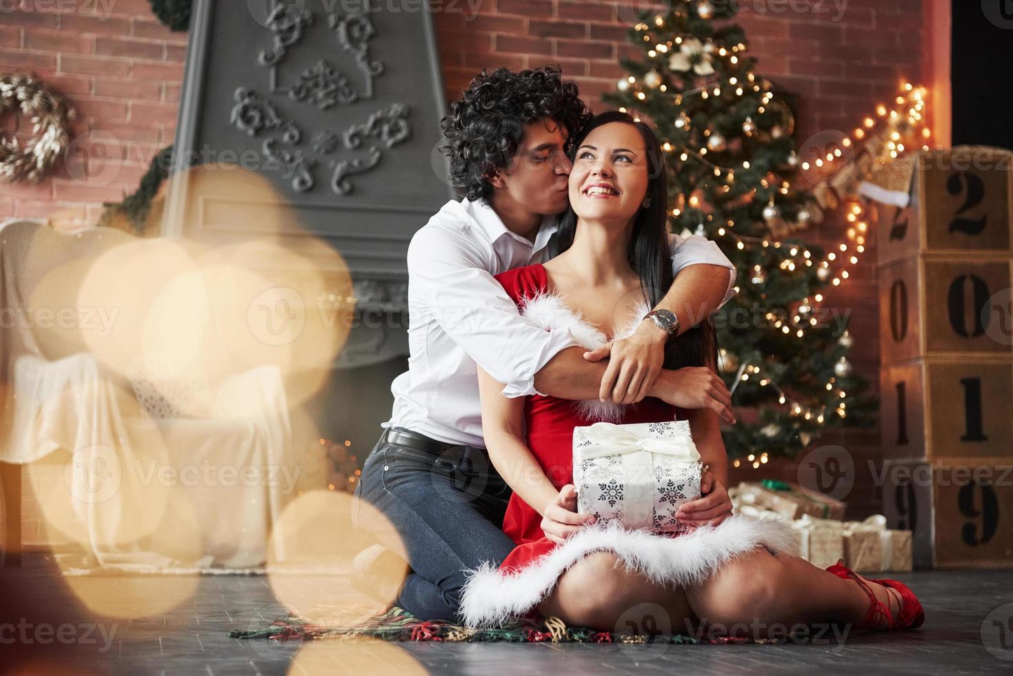 Guy gives a kiss for his woman. Beautiful couple celebrating New year in the decorated room with Christmas tree and fireplace behind photo