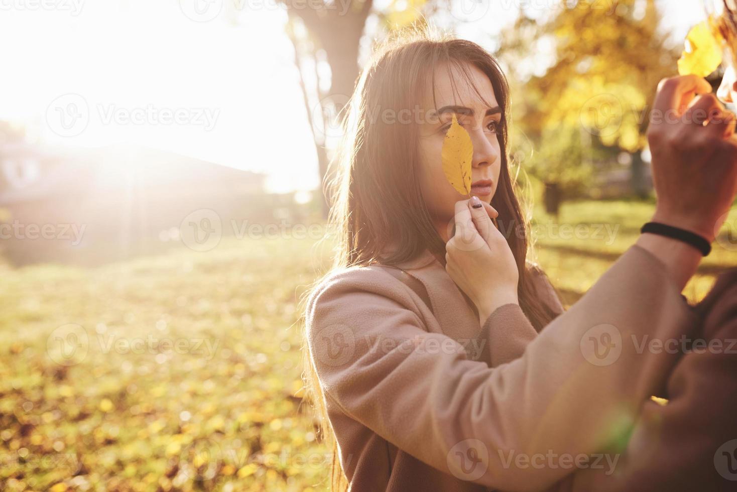 Cerrar retrato de dos bonitas, jóvenes gemelas morenas posando y cubriendo uno de los ojos con una hoja en un abrigo informal en un parque soleado de otoño en el fondo borroso foto