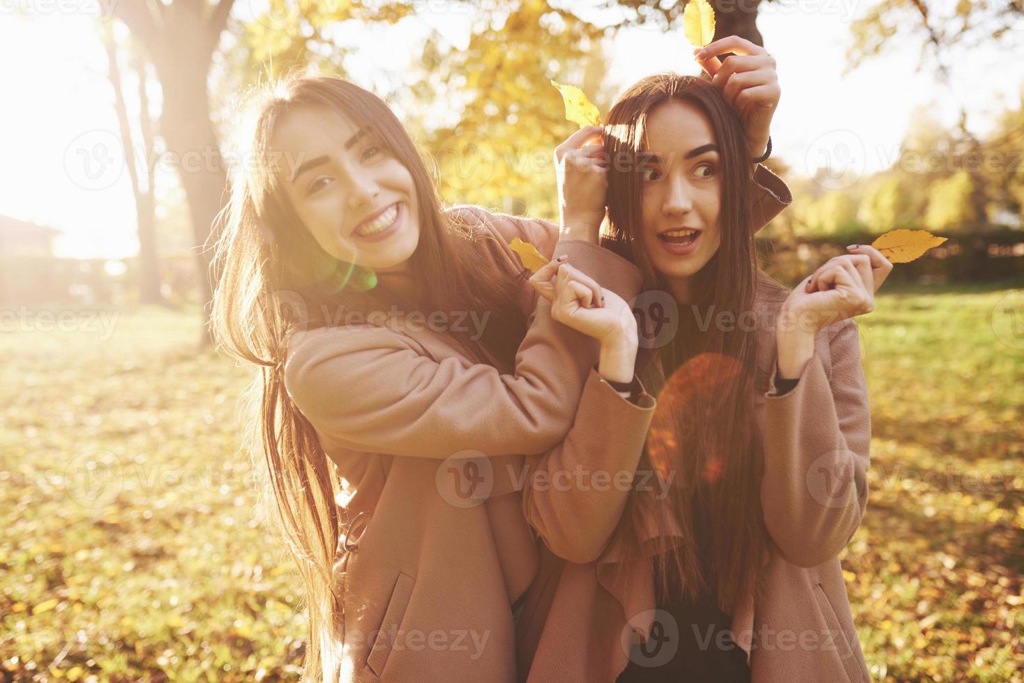 Chicas gemelas morenas jóvenes, bonitas y sonrientes posando, divirtiéndose y jugando con hojas, mientras usan un abrigo informal y están de pie en el parque soleado de otoño sobre fondo borroso foto