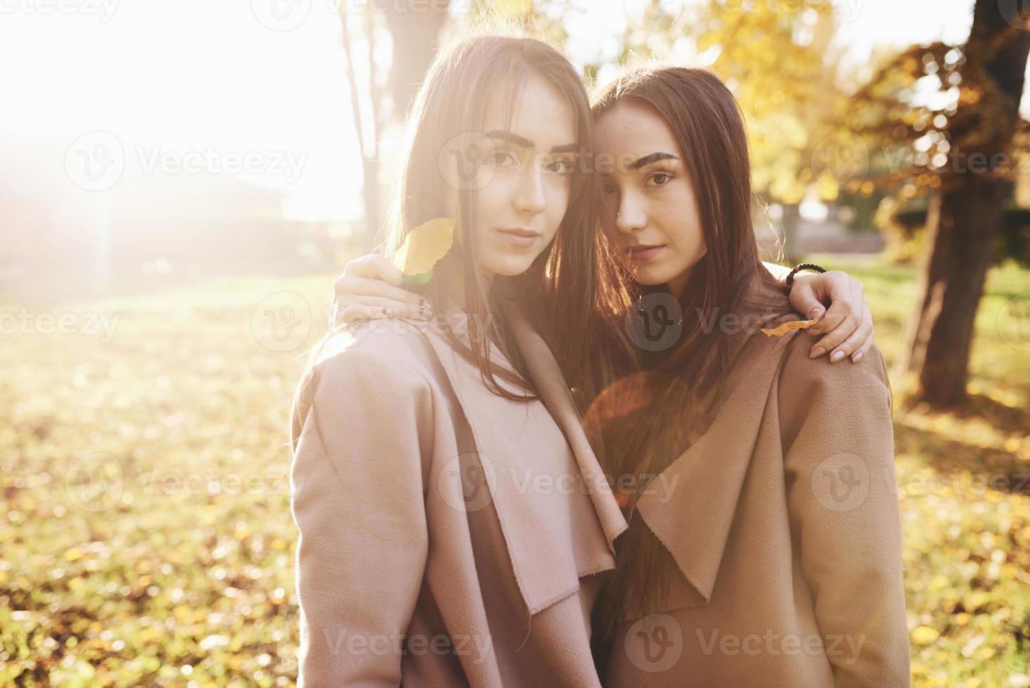 Las jóvenes gemelas morenas de pie muy cerca se volvieron la una a la otra, tomados de la mano con hojas sobre los hombros, vistiendo un abrigo informal, tocándose las cabezas en el parque soleado de otoño sobre fondo borroso foto