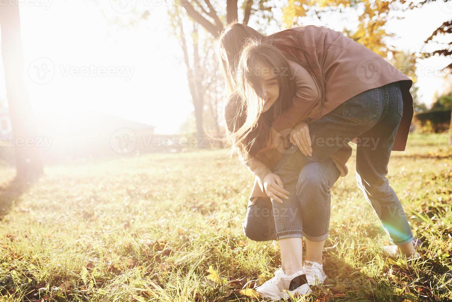 Side profile of young smiling brunette twin girls having fun and wrestling in autumn sunny park on blurry background photo