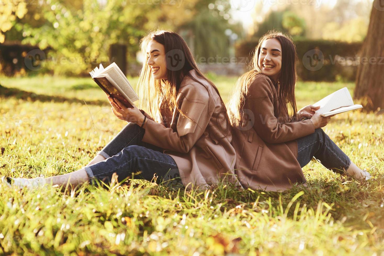 Laughinh brunette twin girls sitting back to back on the grass and having fun with legs slightly bent in knees, with brown books in hands, wearing casual coat in autumn park on blurry background photo