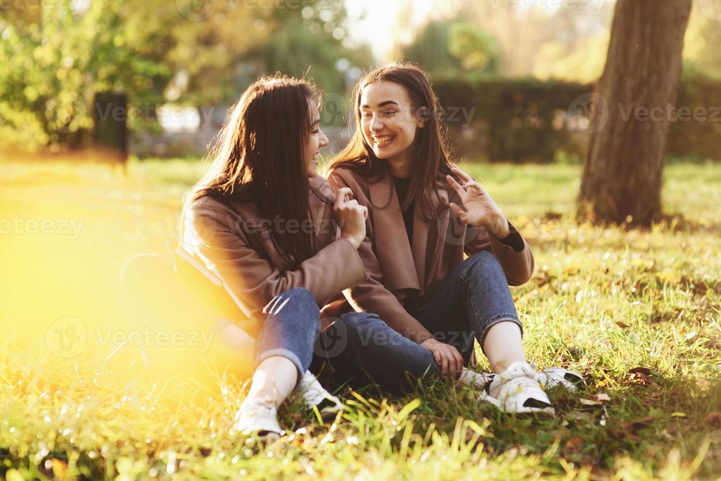 Young smiling brunette twin girls sitting on grass with legs crossed and slightly bent in knees wearing casual coat, chatting, looking at each other at autumn sunny park on blurry background photo