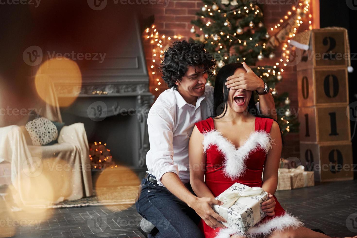 Woman is excited and full of positive emotions. Man making surprise for his girlfriend. Gives New year present. Young couple sits on the floor of room of holiday decorated room photo