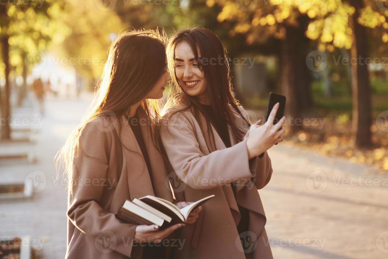 Young smiling brunette twin girls looking at each other and taking selfie with black phone, while one of them is holding books, wearing coat, standing at autumn sunny park alley on blurry background photo