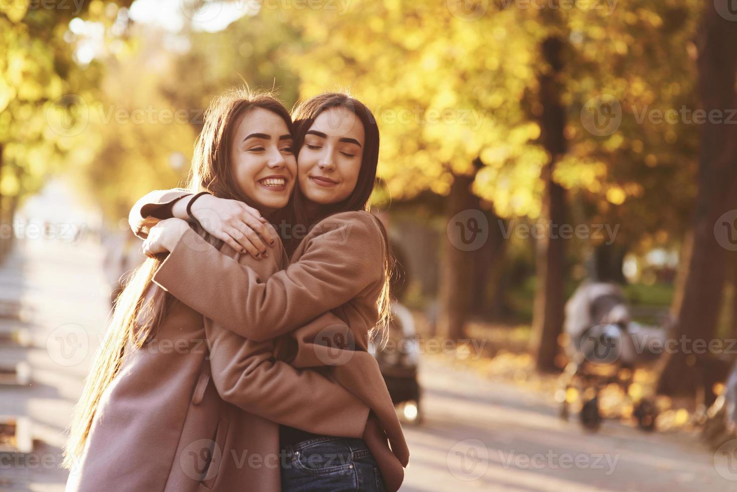 Side profile of young smiling brunette twin girls hugging and having fun in casual coat standing close to each other at autumn sunny park alley on blurry background photo