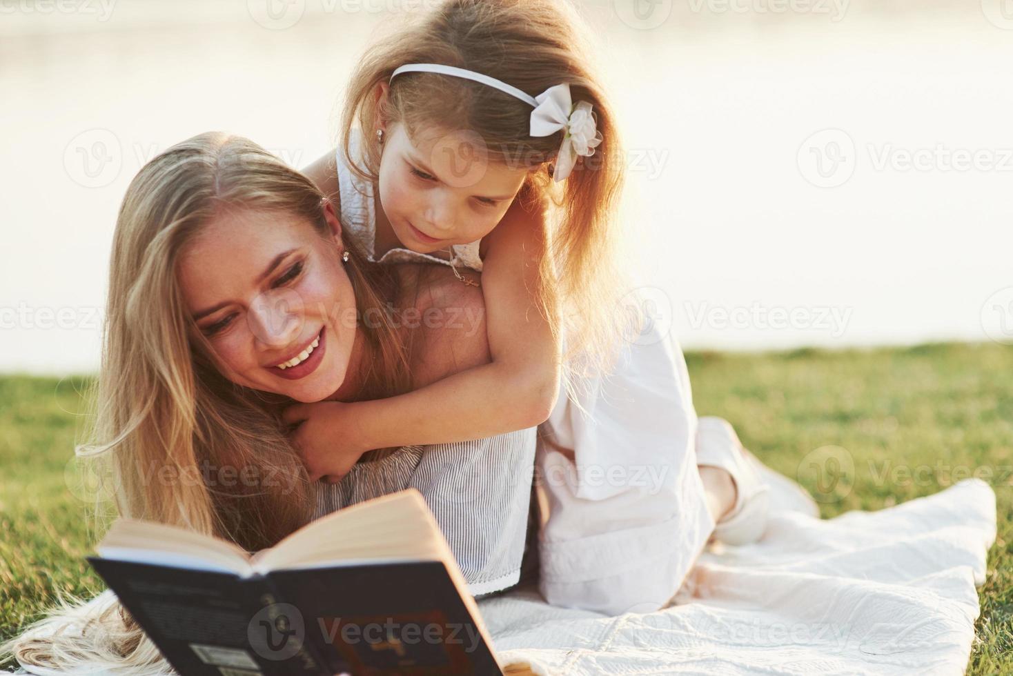 capta una atención muy especial. Mamá e hija leyendo un libro en un día soleado tendido en el césped con el lago al fondo foto