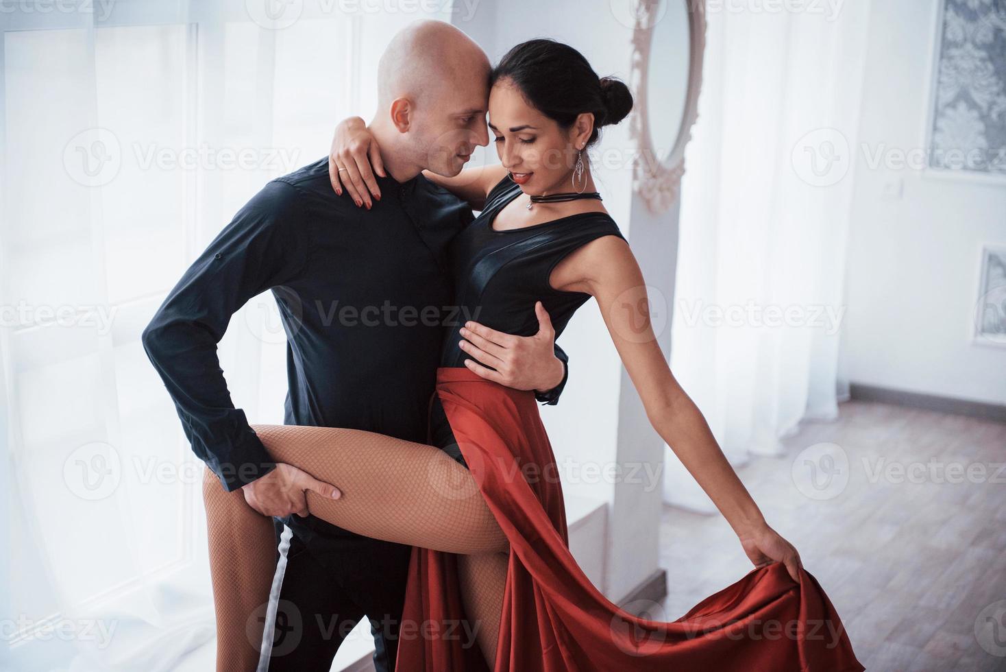 hermosa pose romántica. Joven bonita a mujer vestida de rojo y negro bailando con calvo en la sala blanca foto