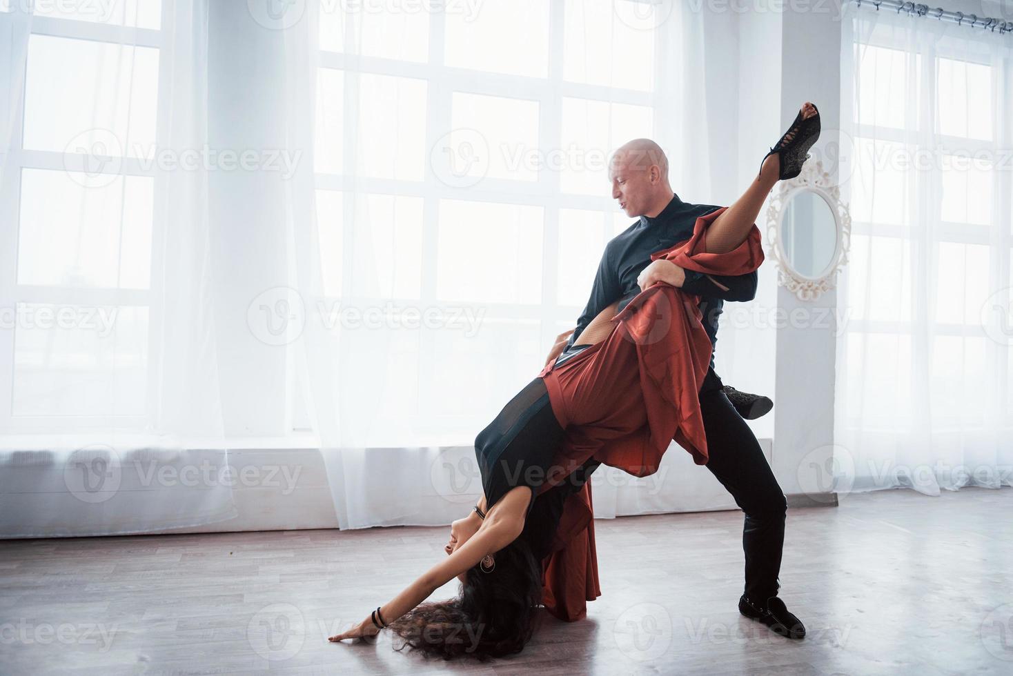 la chica está casi al revés. Joven bonita a mujer vestida de rojo y negro bailando con calvo en la sala blanca foto