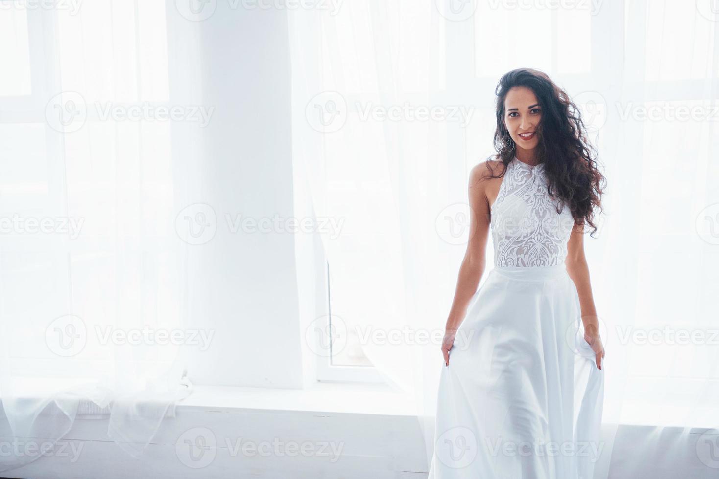 ropa de lujo. Bella mujer vestida de blanco se encuentra en una habitación blanca con luz natural a través de las ventanas foto