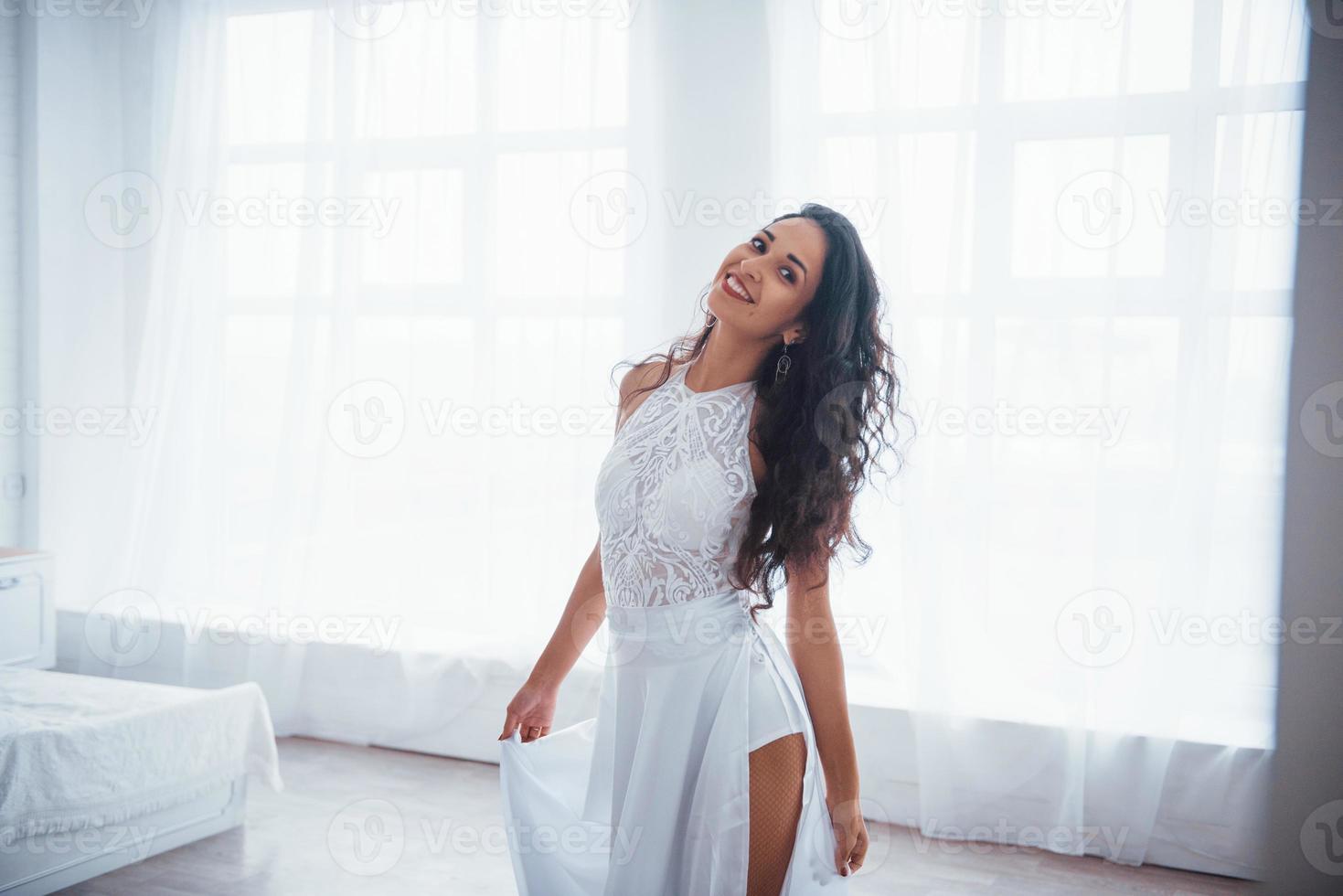 satisfecho y libre. Bella mujer vestida de blanco se encuentra en una habitación blanca con luz natural a través de las ventanas foto