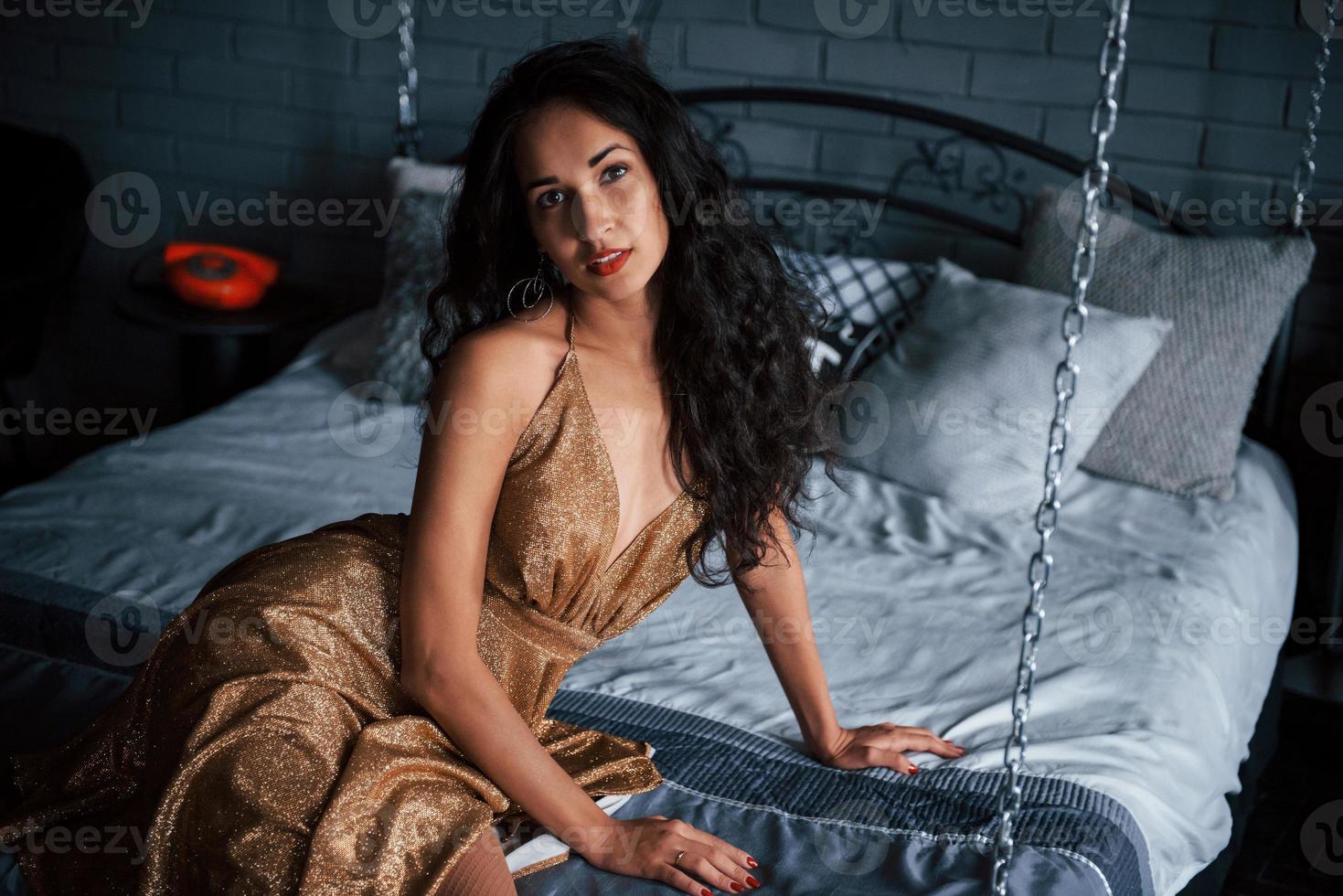 Elegant woman. Girl in golden dress sits at white bed on the chains in luxury apartments photo