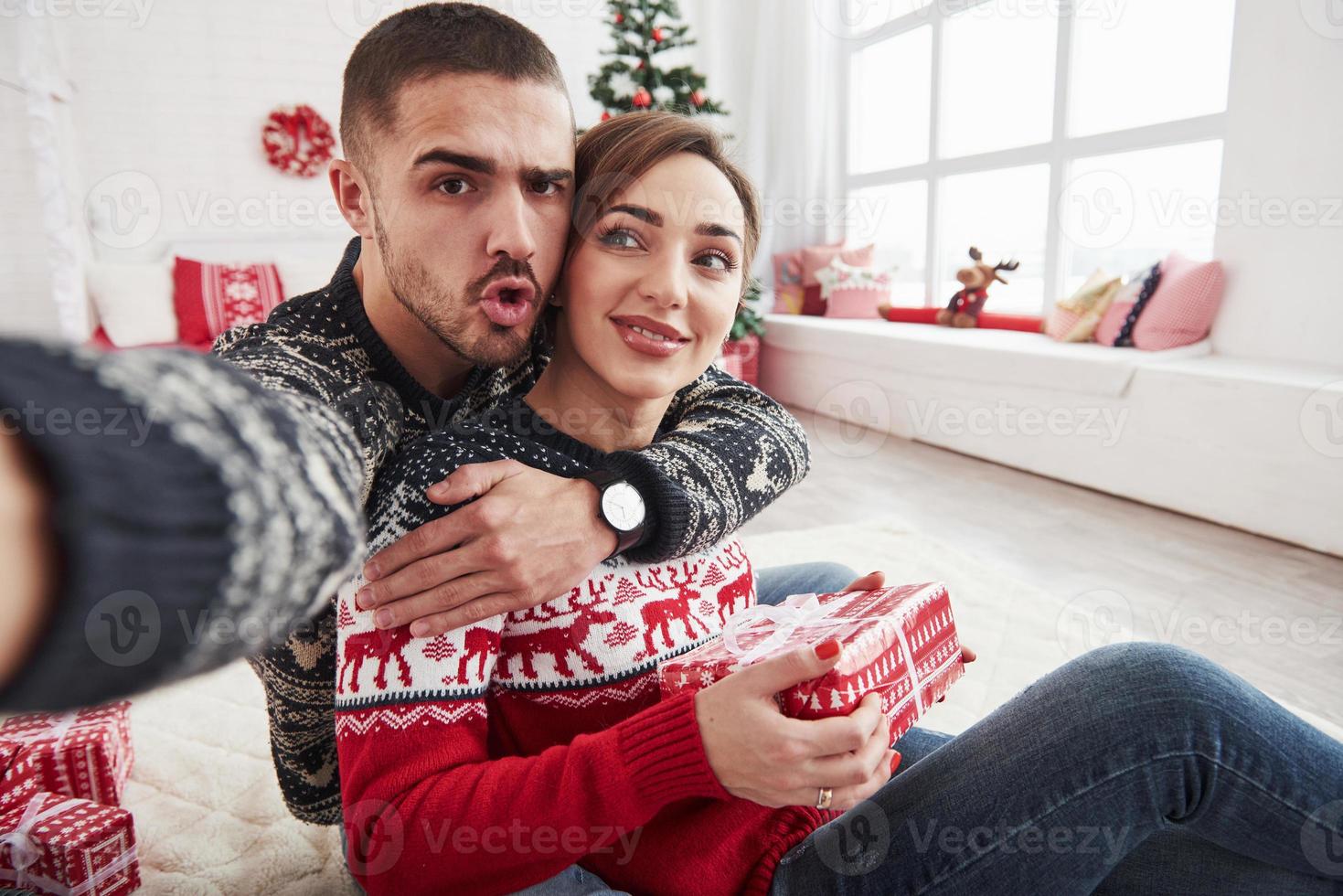chico mira a la cámara. Hombre tomando selfie de él y su esposa vestidos con ropa navideña y sentados en el piso de la hermosa habitación decorativa foto