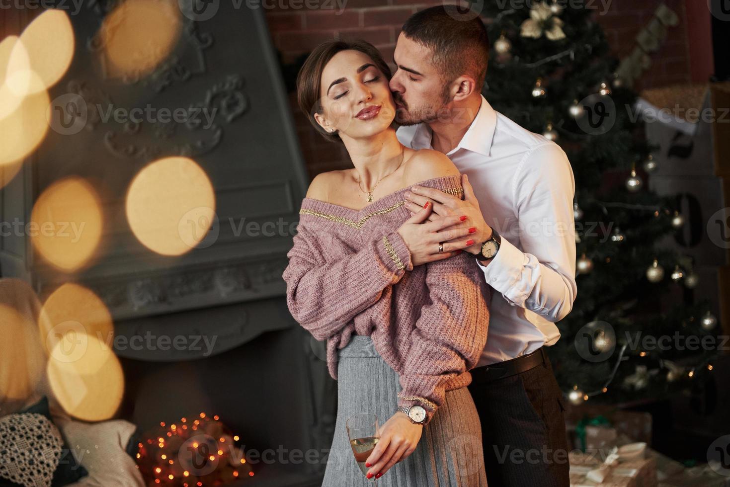 chico besa a su amada chica. Bonita pareja sostiene una copa con champán y celebrando el año nuevo frente al árbol de navidad foto