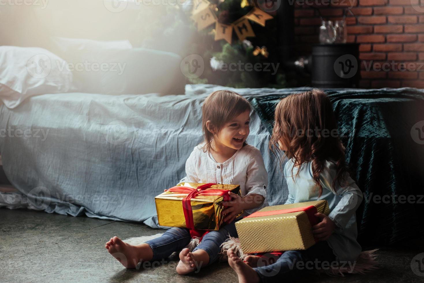 vista frontal. vacaciones navideñas con regalos para estos dos niños que se sientan en el interior de la bonita habitación cerca de la cama foto