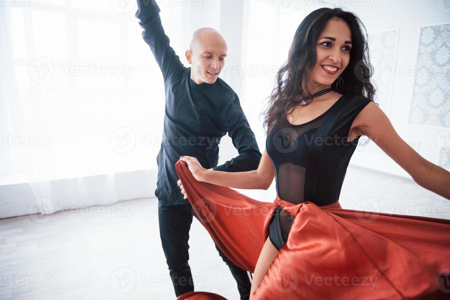 girando y sonriendo. Joven bonita a mujer vestida de rojo y negro bailando con calvo en la sala blanca foto