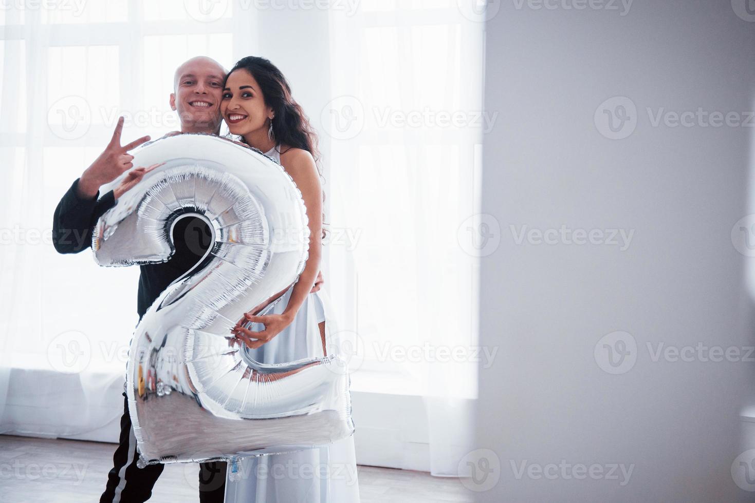 Holding silver colored balloon in the shape on number two. Young couple in luxury wear stands in the white room photo