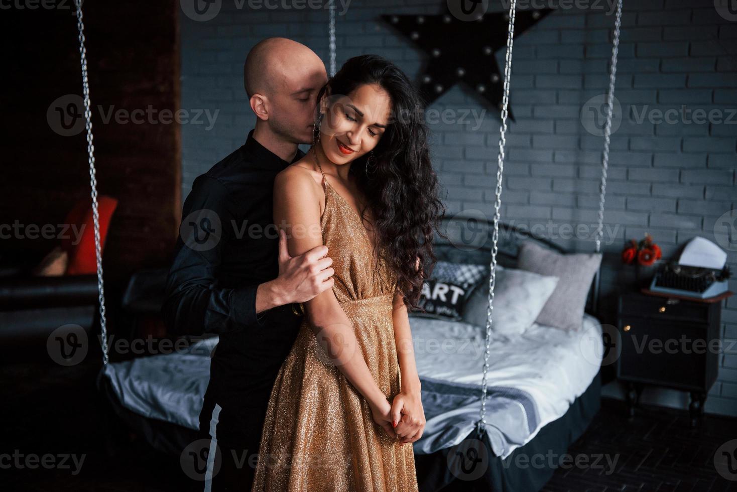 Guy touches girl from the back. Couple in classic wear stands in front of the beautiful luxury decorated bed photo