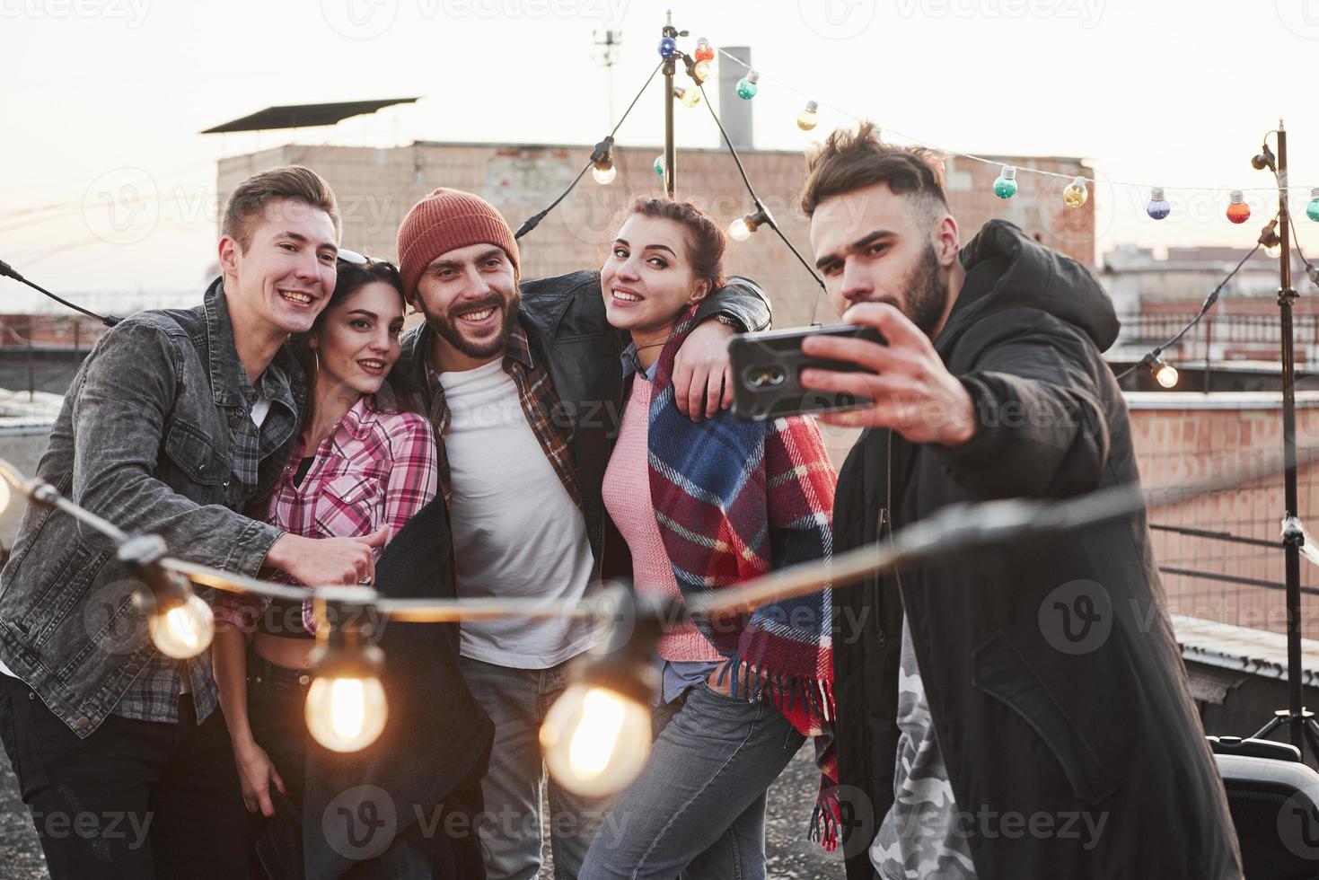 Group of young cheerful friends having fun, hug each other and takes selfie on the roof with decorate light bulbs photo