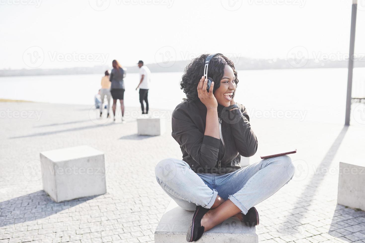Retrato de una hermosa joven bastante afroamericana sentada en la playa o el lago y escuchando música en sus auriculares foto