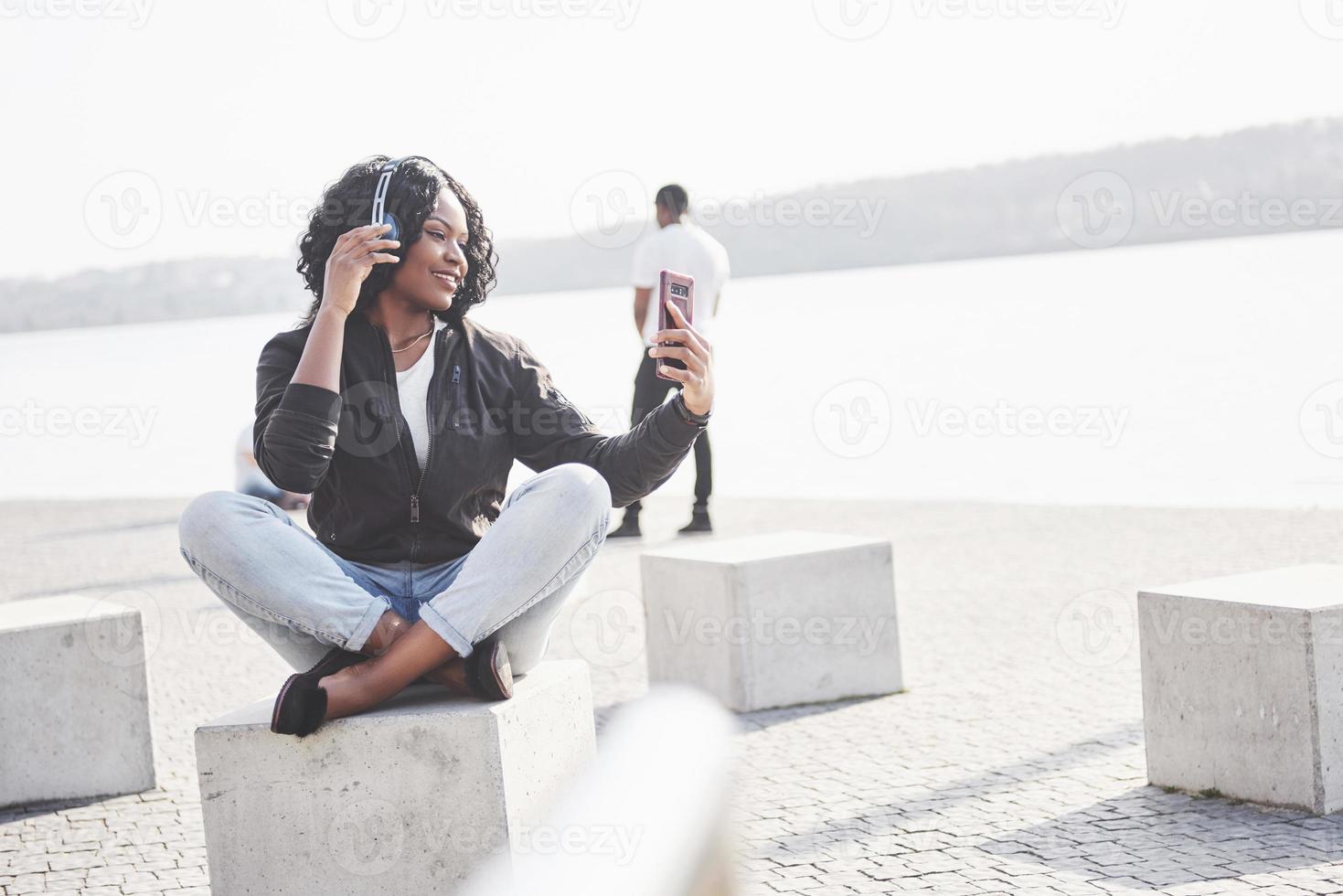 Retrato de una hermosa joven bastante afroamericana sentada en la playa o el lago y escuchando música en sus auriculares foto