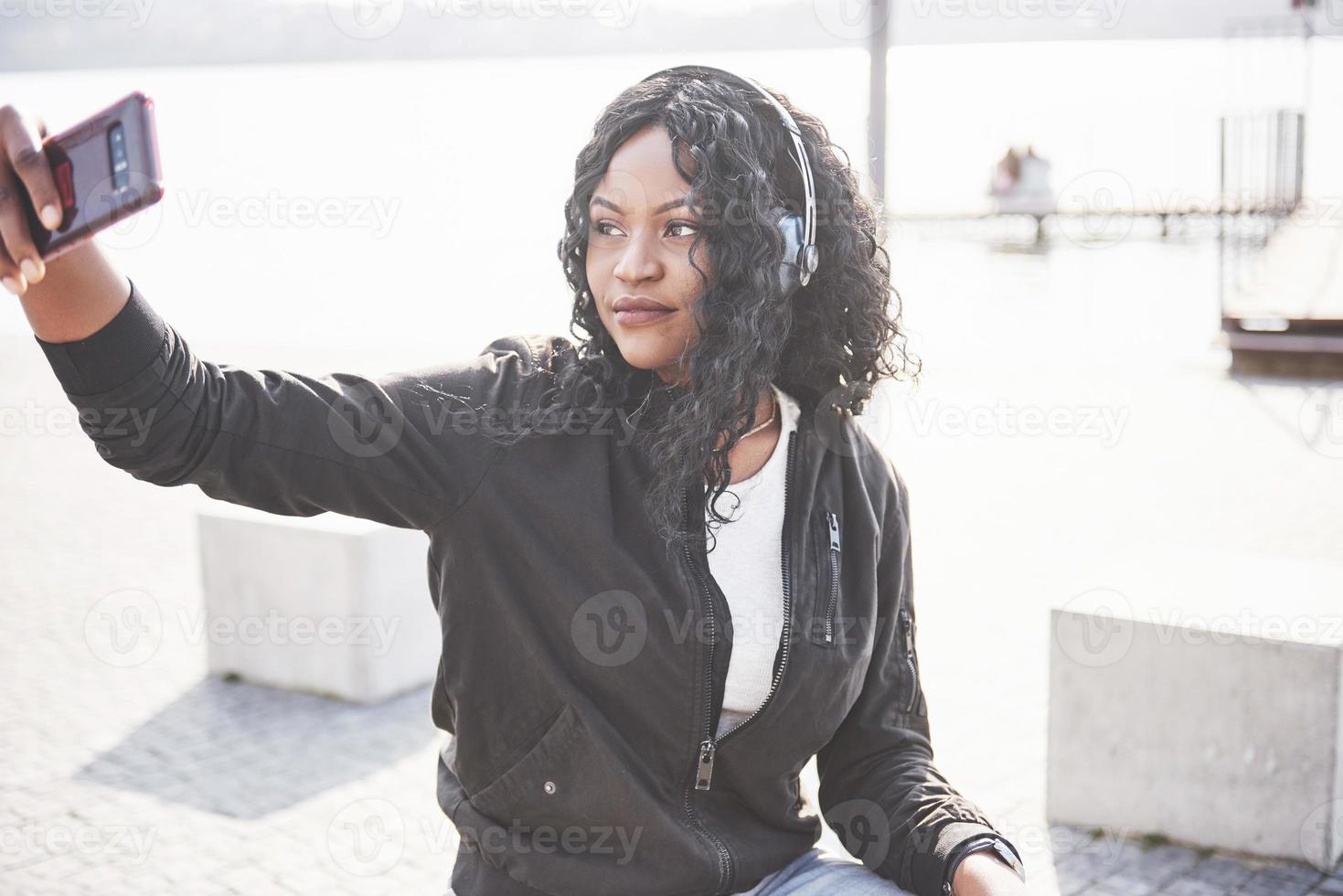 Retrato de una hermosa joven bastante afroamericana sentada en la playa o el lago y escuchando música en sus auriculares foto