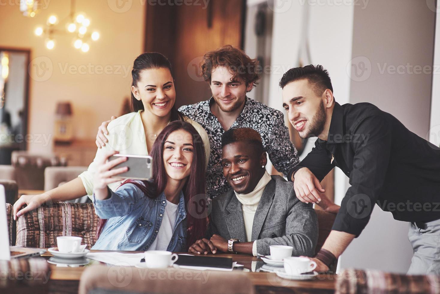 Friends having fun at restaurant. Three boys and two girls making selfie and laughing. On foreground boy holding smart phone. All wear casual clothes photo