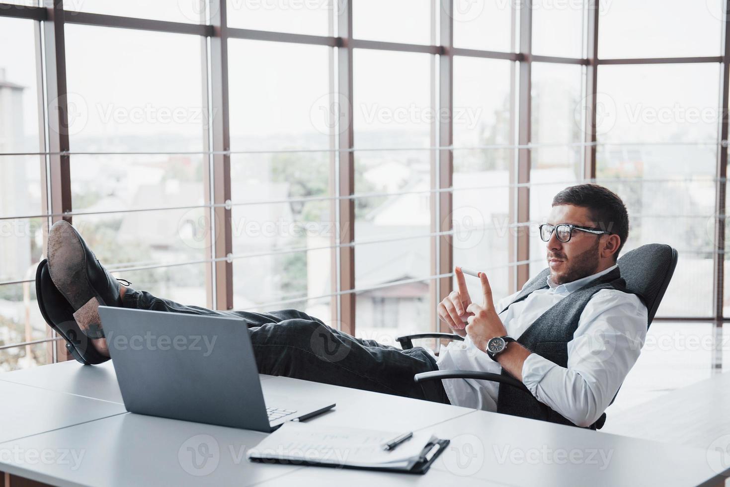 Hermoso joven empresario con gafas sosteniendo sus piernas sobre la mesa mirando una computadora portátil en la oficina foto