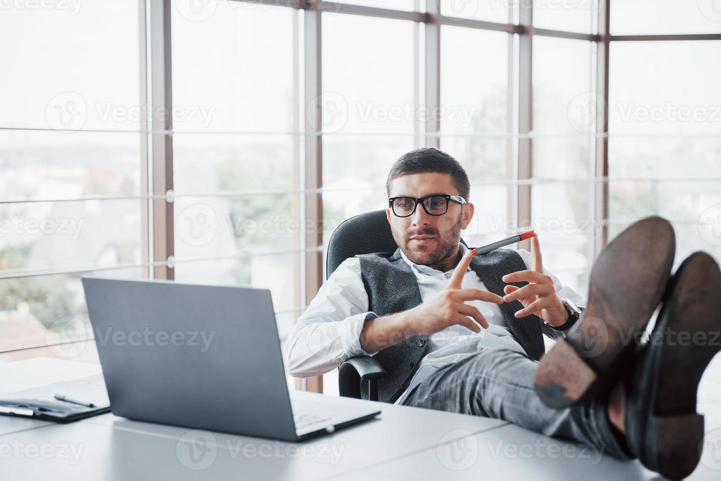 Hermoso joven empresario con gafas sosteniendo sus piernas sobre la mesa mirando una computadora portátil en la oficina foto