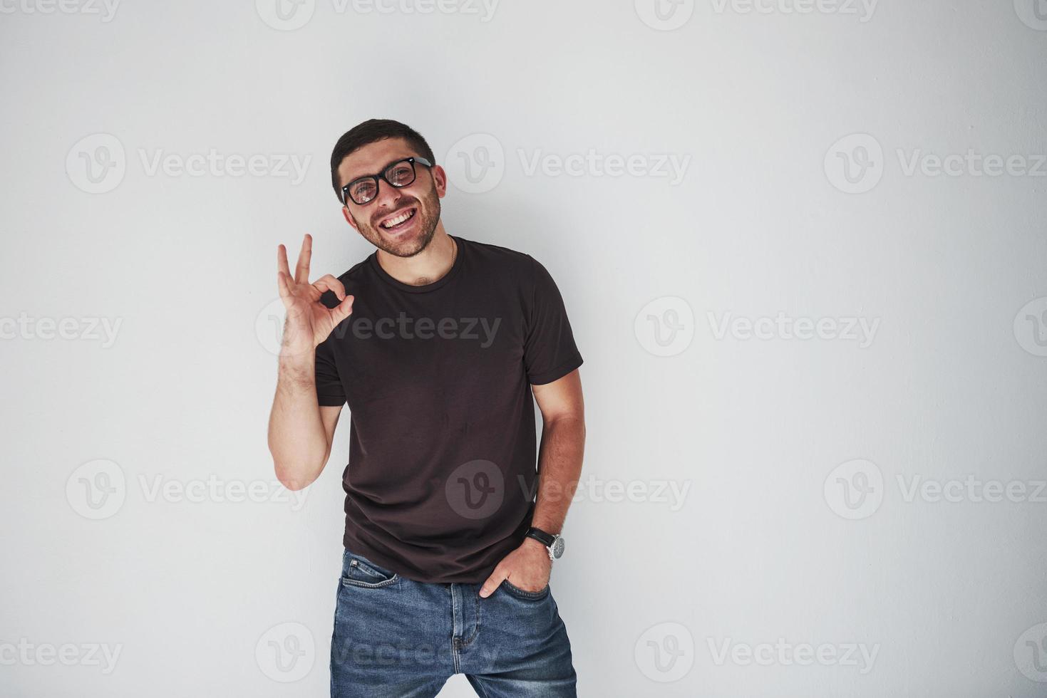 Retrato de un hombre alegre en camiseta y anteojos y mostrando el signo de ok mientras mira a la cámara sobre fondo blanco. foto