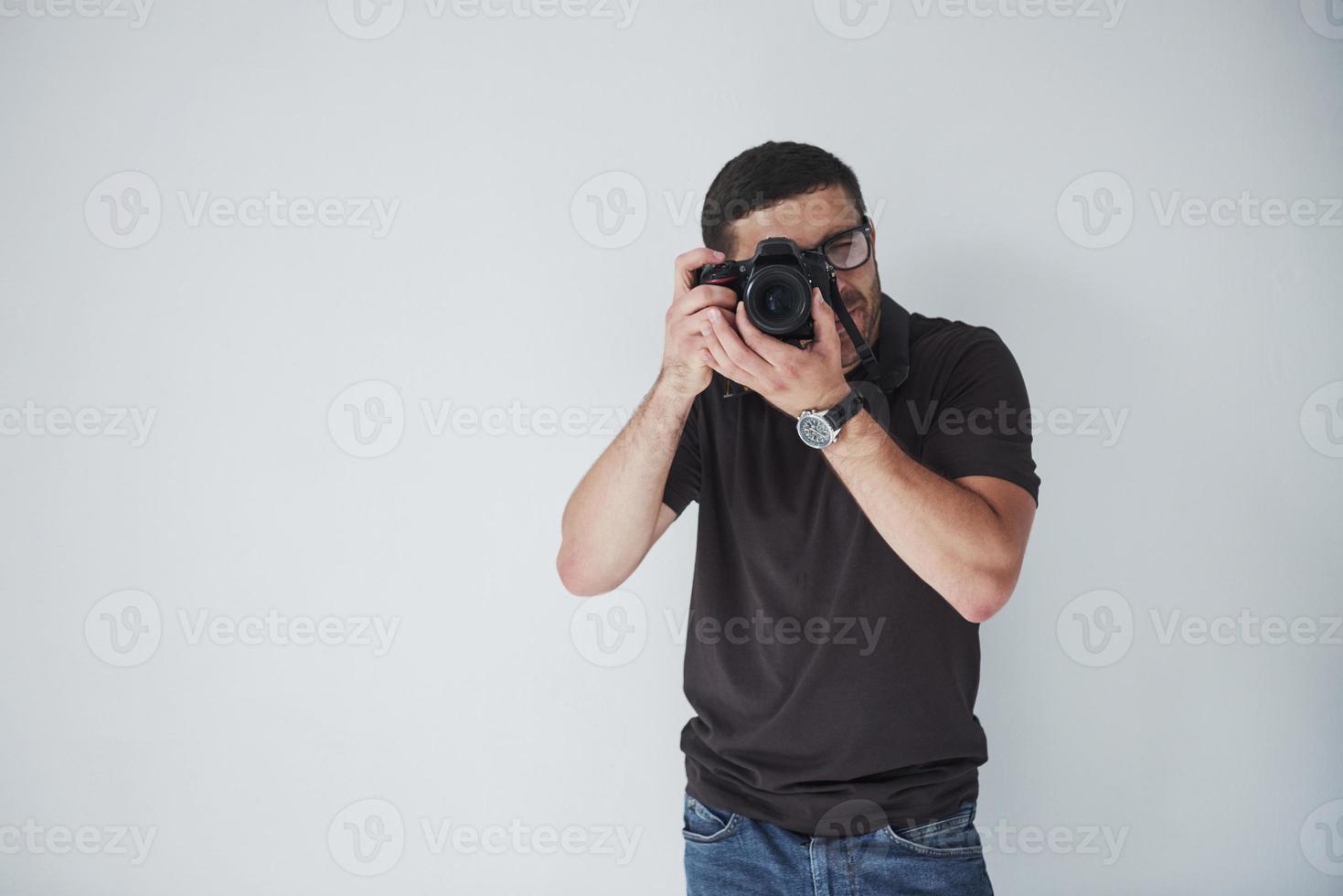 A young hipster man in eyepieces holds a DSLR camera in hands standing against a white wall background photo