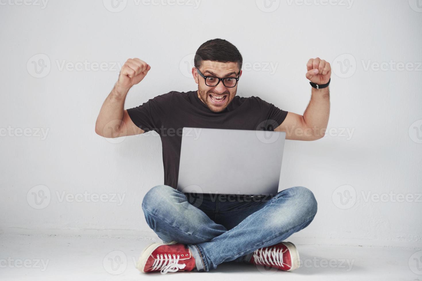 Joven hombre caucásico sentado sobre una pared de ladrillo blanco con ordenador portátil feliz en estado de shock con una cara de sorpresa, asustado y emocionado con expresión de miedo foto