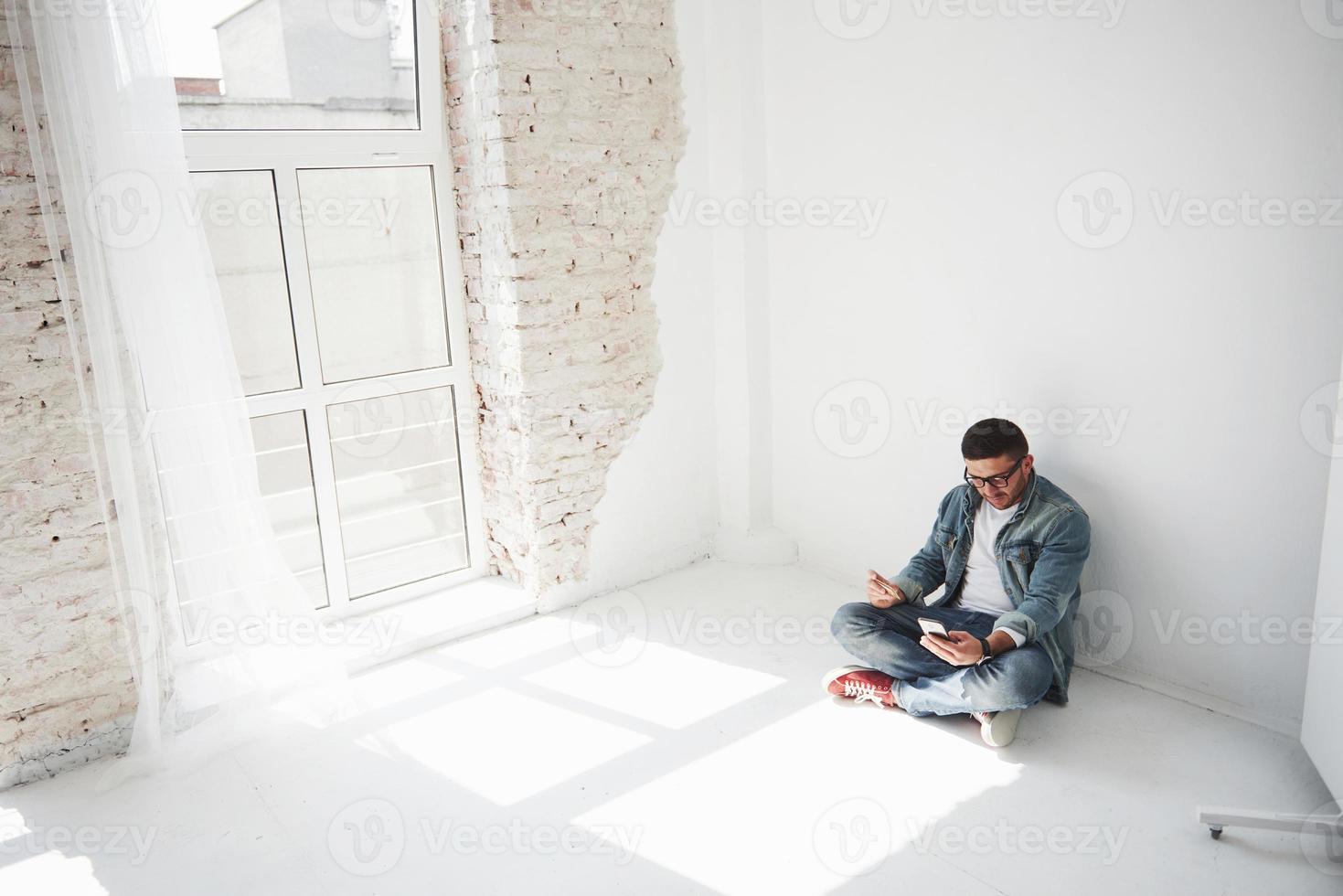 un chico con ropa informal está sentado en su casa en un apartamento vacío con una tarjeta de crédito y llama por teléfono. tal vez sea un residente nuevo y aún no haya comprado ningún mueble. foto