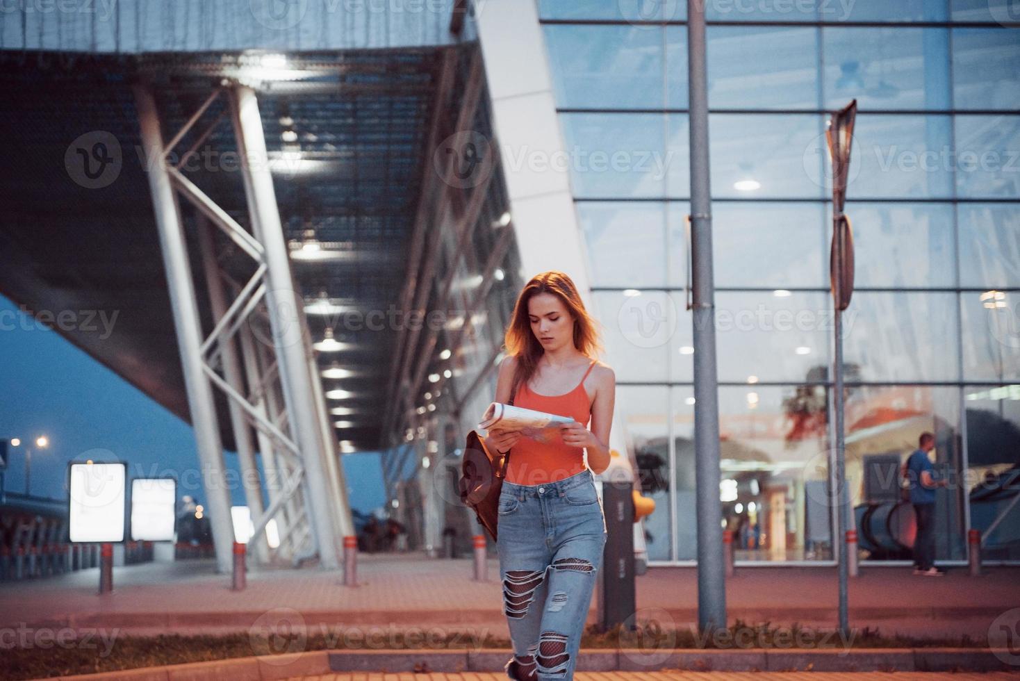 Young girl costs at night near the terminal of the airport or station and reading city map and looking for hotel. Cute tourist with backpacks determine the concept of travel photo