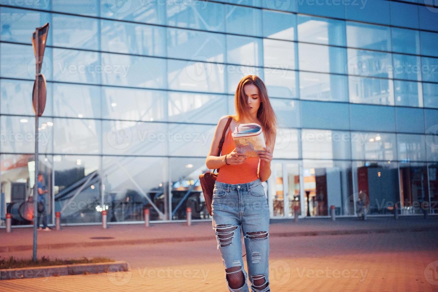 Young girl costs at night near the terminal of the airport or station and reading city map and looking for hotel. Cute tourist with backpacks determine the concept of travel photo