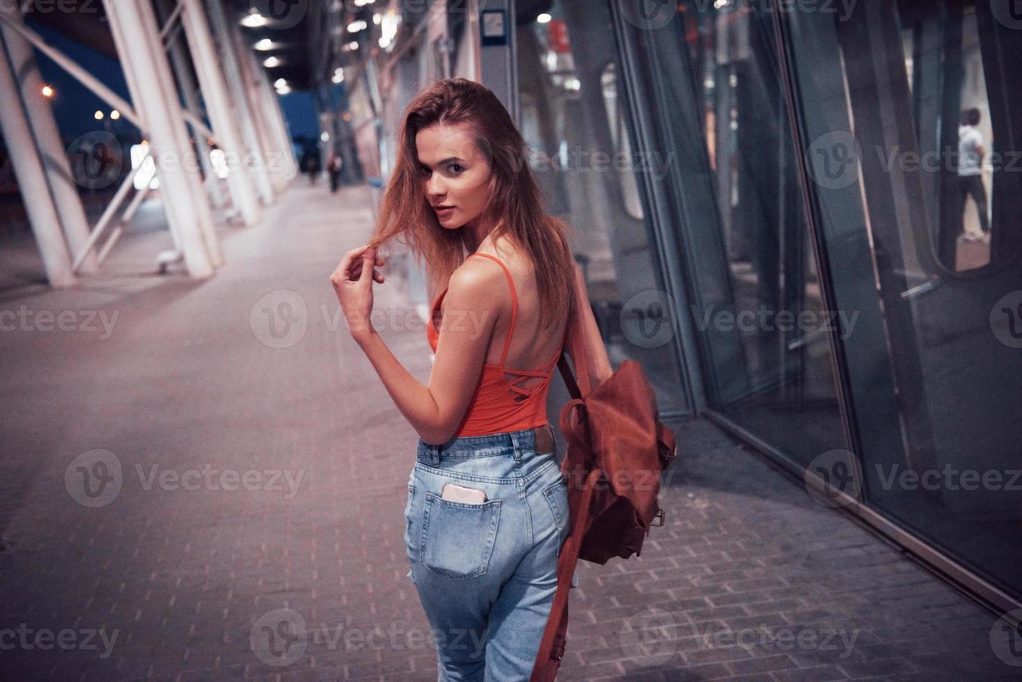 una hermosa joven con una mochila detrás de los hombros de pie en la calle cerca de un aeropuerto. ella acaba de llegar del resto y muy feliz foto