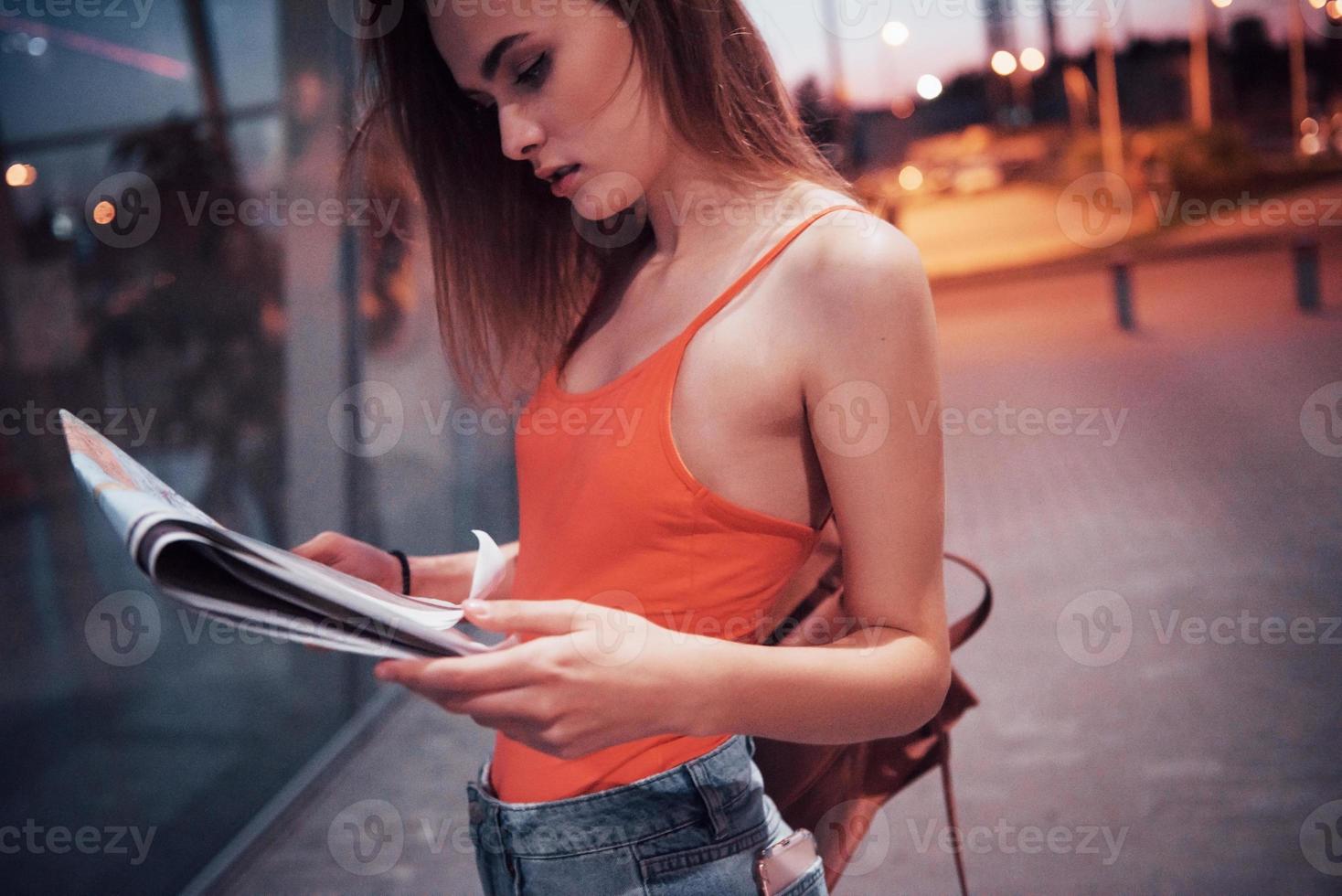 Young girl costs at night near the terminal of the airport or station and reading city map and looking for hotel. Cute tourist with backpacks determine the concept of travel photo