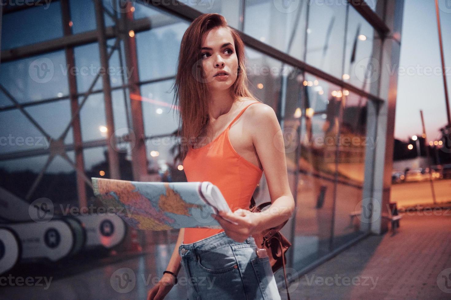 Young girl costs at night near the terminal of the airport or station and reading city map and looking for hotel. Cute tourist with backpacks determine the concept of travel photo