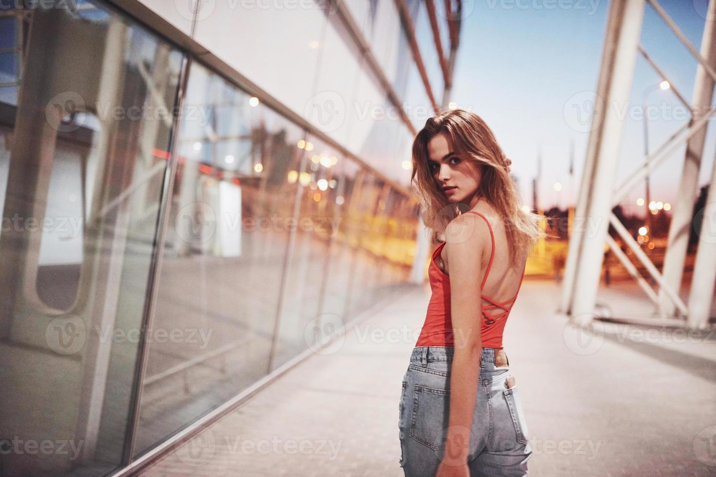 hermosa chica sexy turista caminando en la concurrida calle de la ciudad cerca del aeropuerto. Mujer mirando a la cámara al aire libre vistiendo una camiseta roja de moda foto