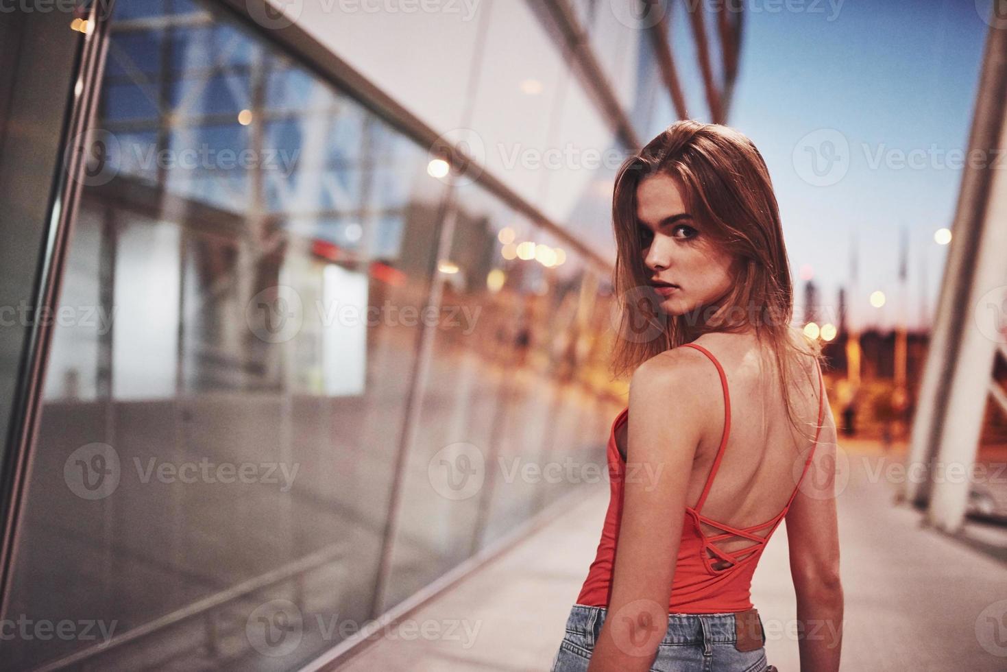 hermosa chica sexy turista caminando en la concurrida calle de la ciudad cerca del aeropuerto. Mujer mirando a la cámara al aire libre vistiendo una camiseta roja de moda foto