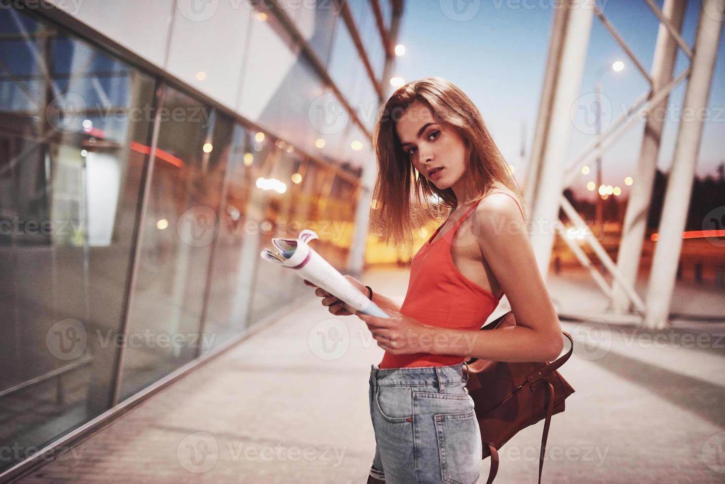 una hermosa joven con una mochila detrás de los hombros de pie en la calle cerca de un aeropuerto. ella acaba de llegar del resto y muy feliz foto