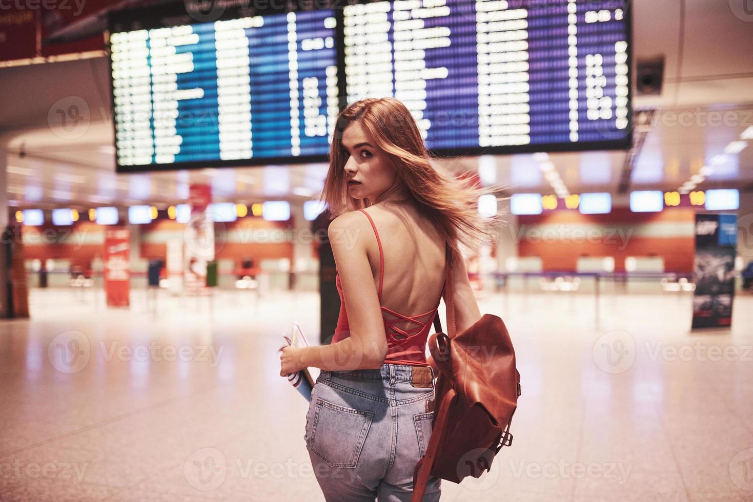 Hermosa joven turista con mochila en el aeropuerto internacional, cerca del tablero de información de vuelos foto